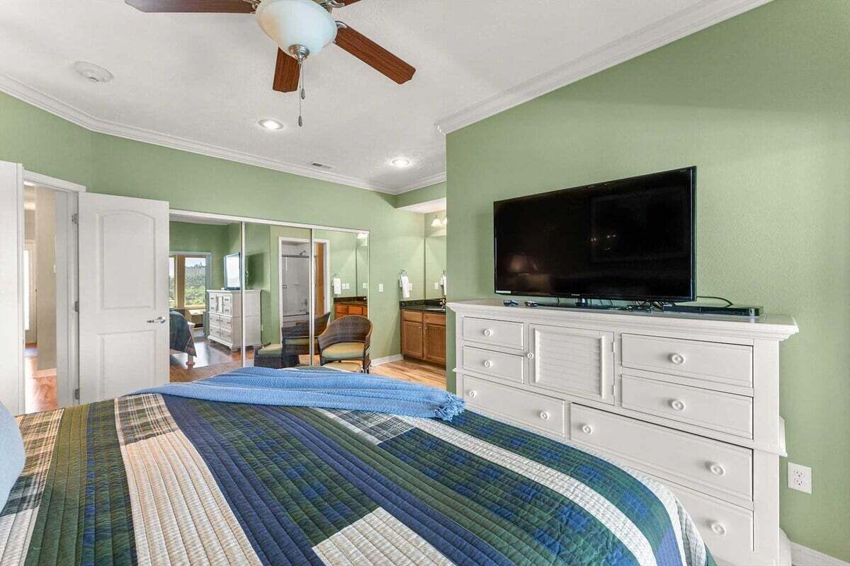 A bedroom with a bed, a television on a dresser, a ceiling fan, and a door leading to another room.