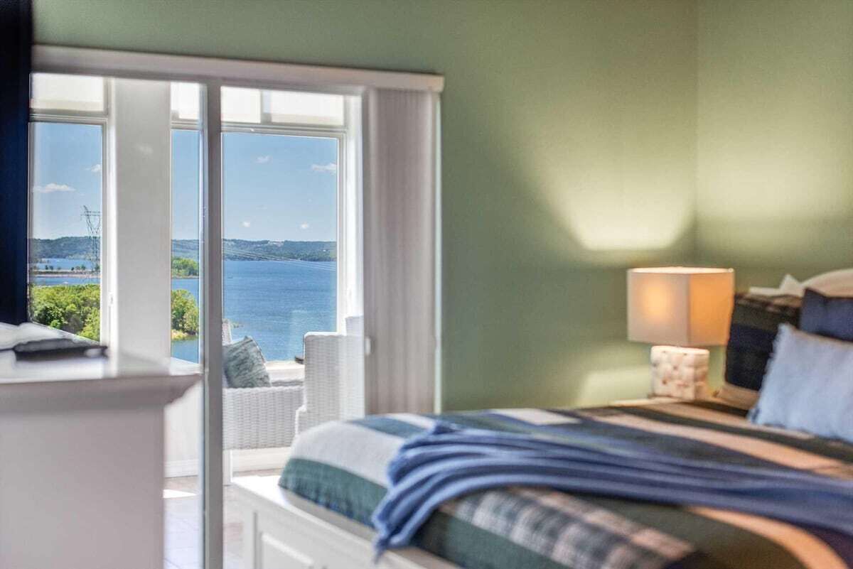 A bedroom with a bed, a nightstand, a lamp, and a view of the sea through large windows.