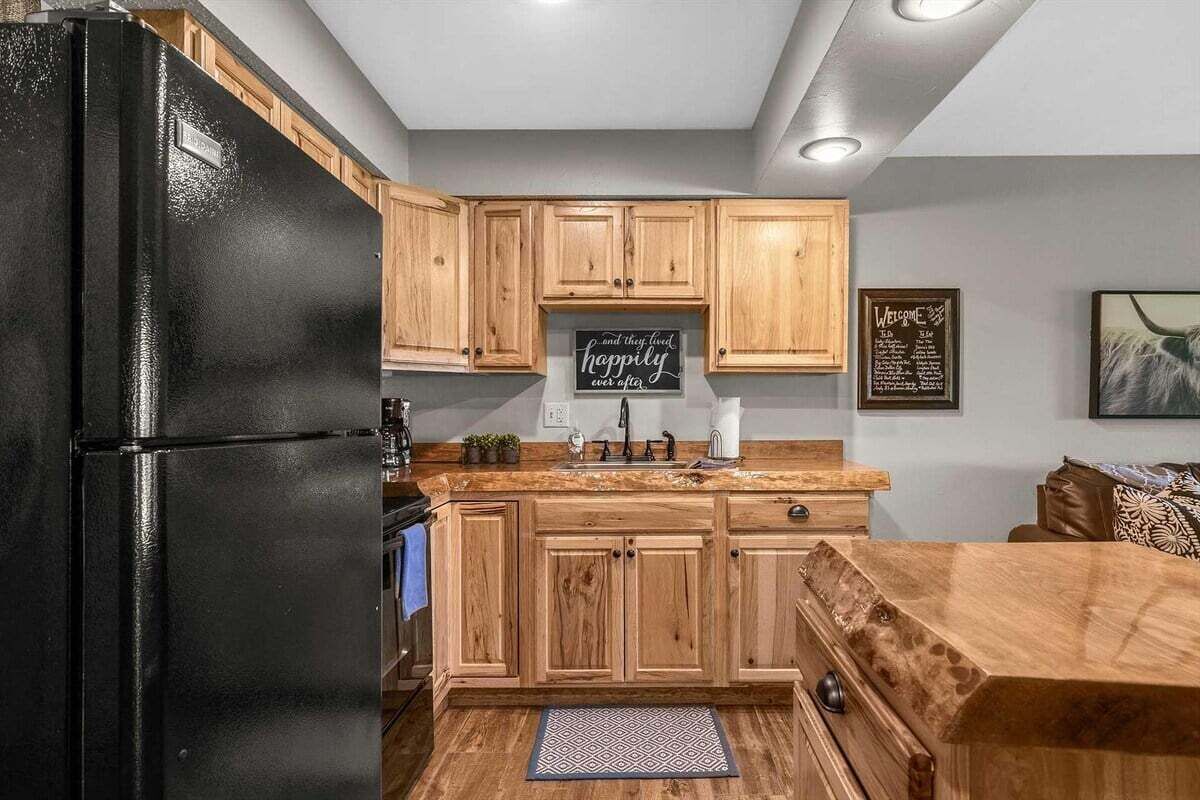 A kitchen with wooden cabinets, a black refrigerator, a chalkboard with a message, and a framed welcome sign.