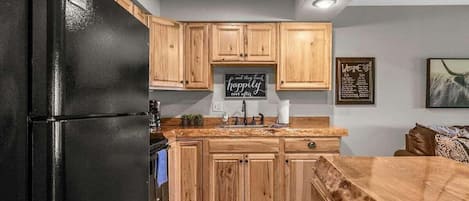 A kitchen with wooden cabinets, a black refrigerator, a chalkboard with a message, and a framed welcome sign.
