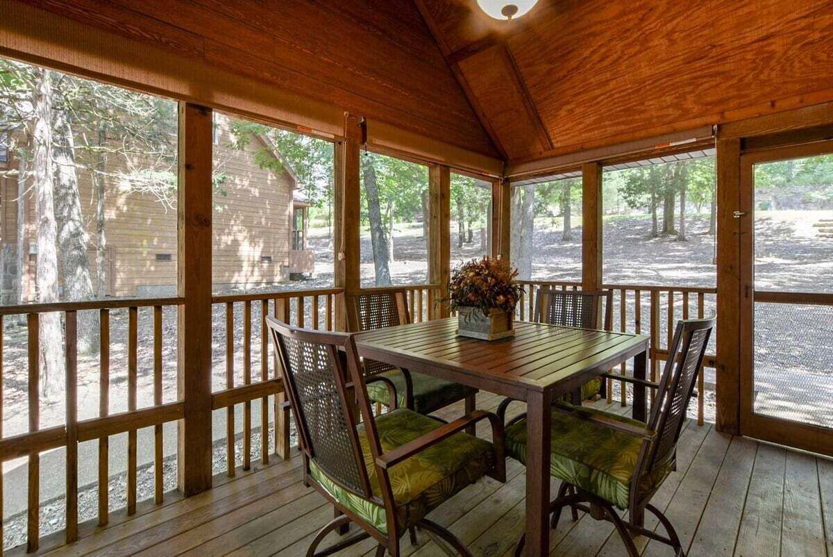 A covered outdoor dining area with a table and chairs, surrounded by wooden railings and large windows.