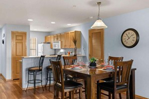 A dining area with a wooden table and chairs, a kitchen in the background, and a wall clock. - Table Rock Condo_Huge Deck_New Flooring_Private_Free Tickets (Hollister)