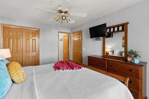 Ein Schlafzimmer mit Bett, Holzschrank, Schminktisch und Deckenventilator, sowie einem an der Wand befestigten Fernseher.