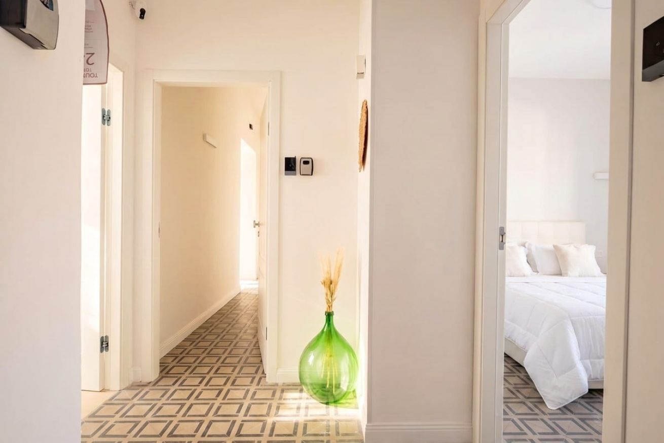 A hotel hallway with a patterned floor, a green vase, and a bed with white bedding in the adjacent room.