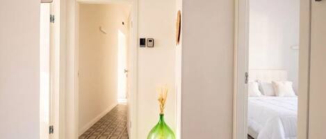 Un pasillo de hotel con piso estampado, un jarrón verde y una cama con ropa blanca en la habitación contigua.