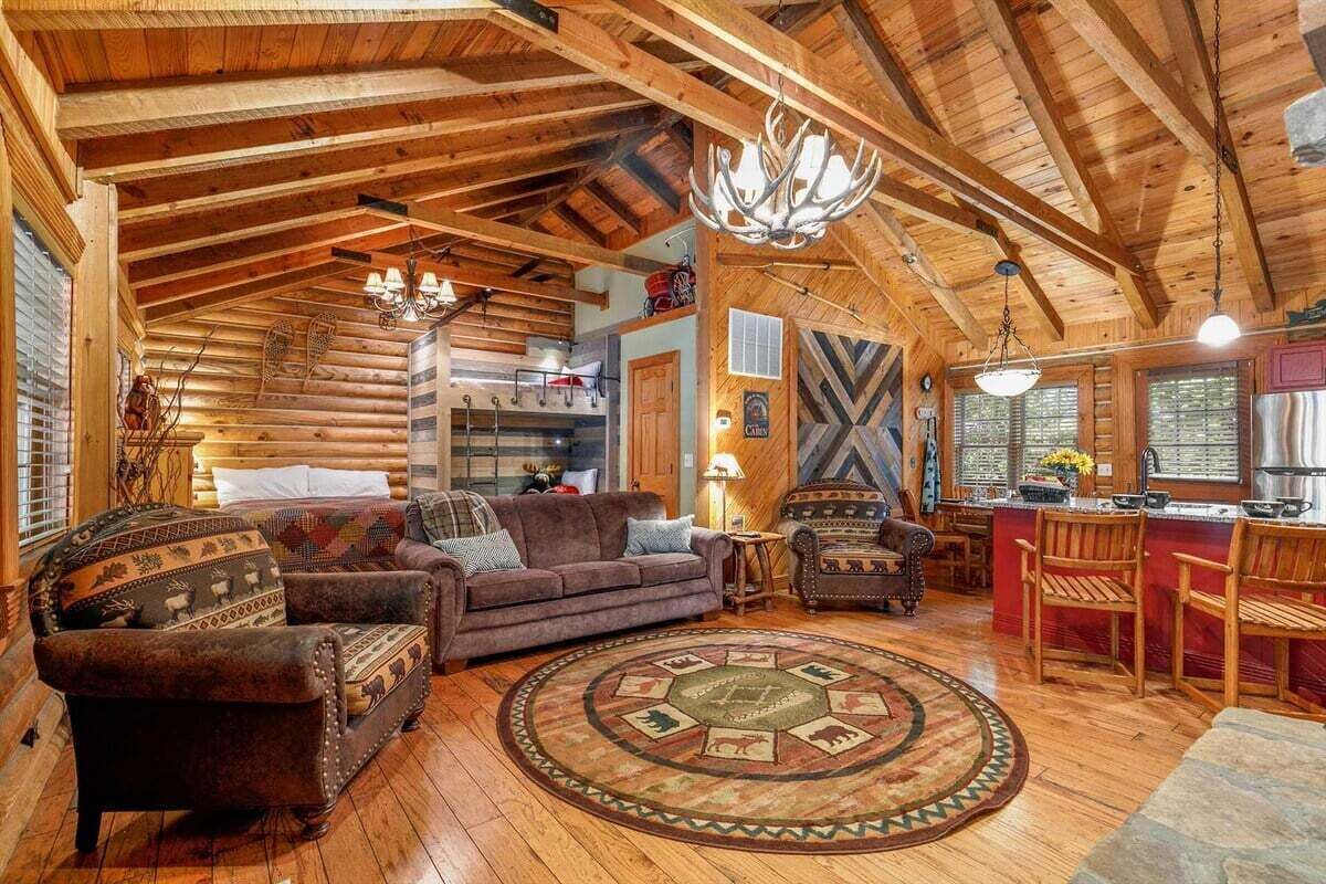 L’intérieur d’une cabane en rondins, avec un plafond en bois, un tapis rond, un canapé, un fauteuil et un coin cuisine.