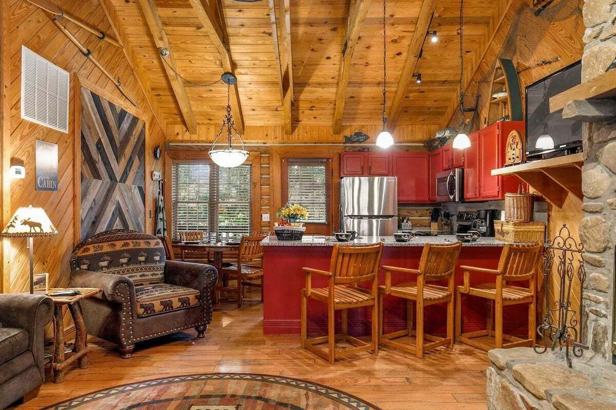 A cabin interior with a wooden ceiling, a fireplace, a kitchen area, and a seating area with a sofa and chairs.