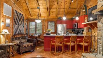 A cabin interior with a wooden ceiling, a fireplace, a kitchen area, and a seating area with a sofa and chairs.