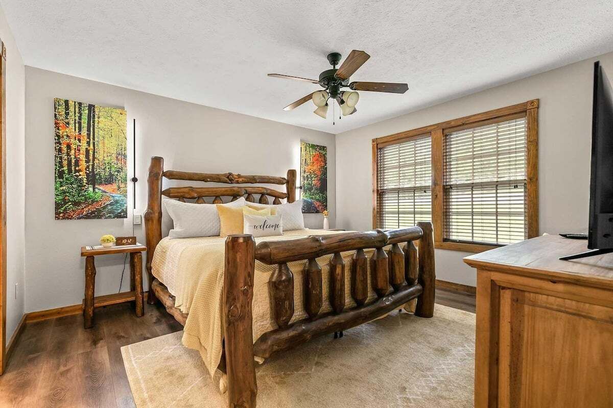 A bedroom with a wooden bed, a desk, and two windows with blinds.
