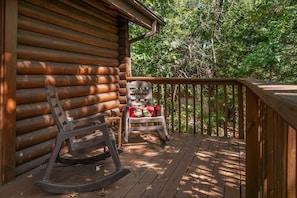 A wooden deck with a rocking chair and a chair with a cushion, surrounded by trees. - Genuine Log Cabin_Soaker Tub_Gas Fireplace_Great Location_Free Daily Attraction Tickets (Branson)