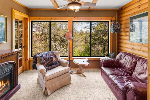 A log cabin interior with a fireplace, a leather couch, a patterned armchair, and large windows offering a view of trees.