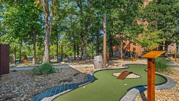 A miniature golf course with a winding path, wooden structures, and a backdrop of trees and buildings.