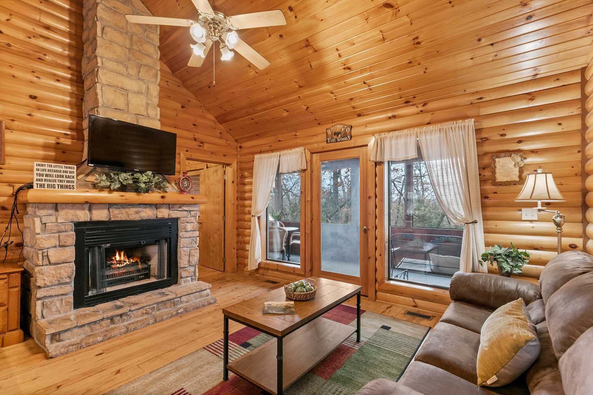 A cozy cabin interior with a fireplace, wooden walls, a ceiling fan, and a sofa.