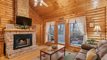 A cozy cabin interior with a fireplace, wooden walls, a ceiling fan, and a sofa.