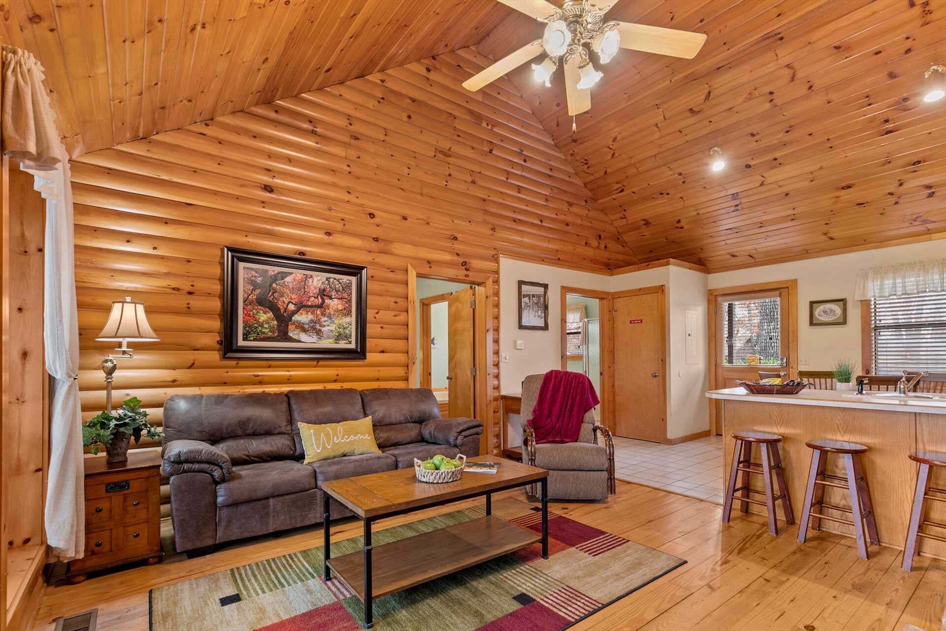A cozy cabin interior with a wooden ceiling, a sofa, a coffee table, a dining area with stools, and a kitchen visible through an open door.