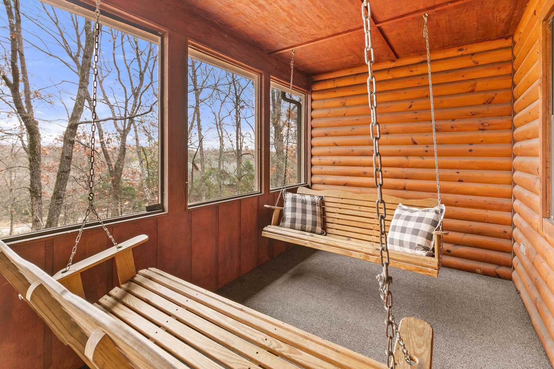L’intérieur d’une cabane en rondins, avec un banc en bois, un siège suspendu avec des coussins et de grandes fenêtres donnant sur la forêt.