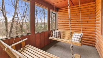 L’intérieur d’une cabane en rondins, avec un banc en bois, un siège suspendu avec des coussins et de grandes fenêtres donnant sur la forêt.
