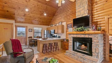 A cozy cabin interior with a stone fireplace, wooden ceiling, and a kitchen area.