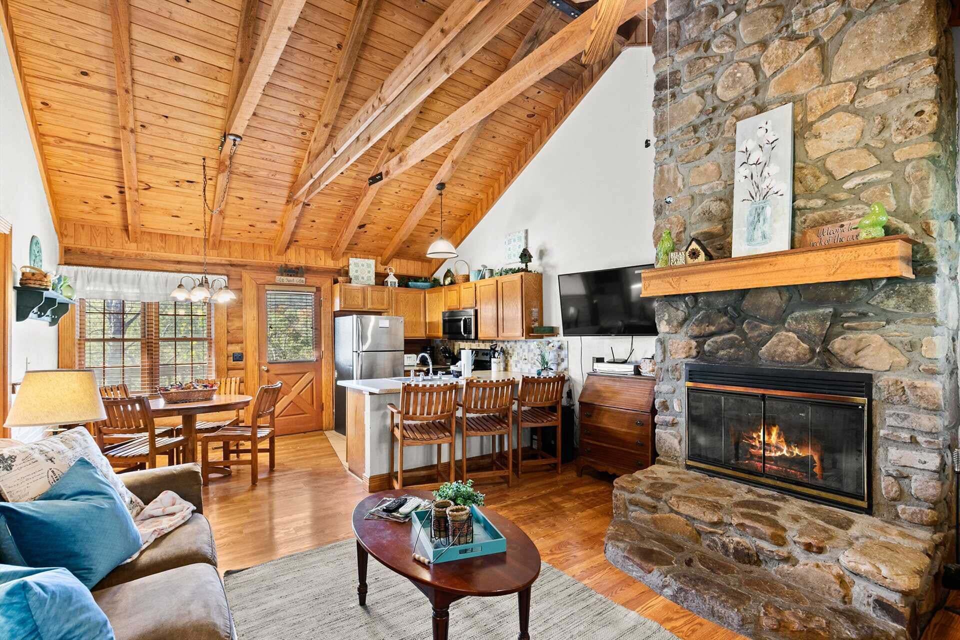 A cozy cabin interior with a stone fireplace, wooden ceiling beams, a kitchen area, and a dining space.