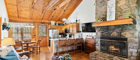 A cozy cabin interior with a stone fireplace, wooden ceiling beams, a kitchen area, and a dining space.