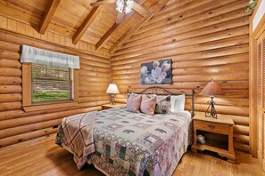 Ein Schlafzimmer in einer Blockhütte mit einem Bett, zwei Nachttischen, einem Fenster mit Jalousien, einem Deckenventilator und einem Bild an der Wand.