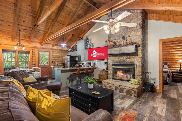 A cozy cabin interior with a stone fireplace, wooden ceiling beams, a dining area, and a kitchen. - Log Cabin_ Resort Amenities_Free Daily Attraction Tickets (Branson)