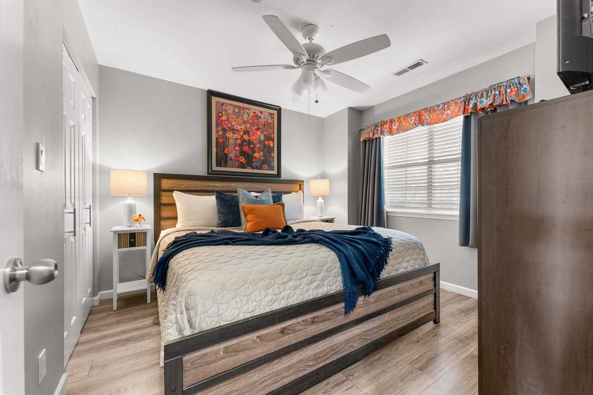 A bedroom with a bed, bedside lamps, a ceiling fan, a painting, and a window with curtains.