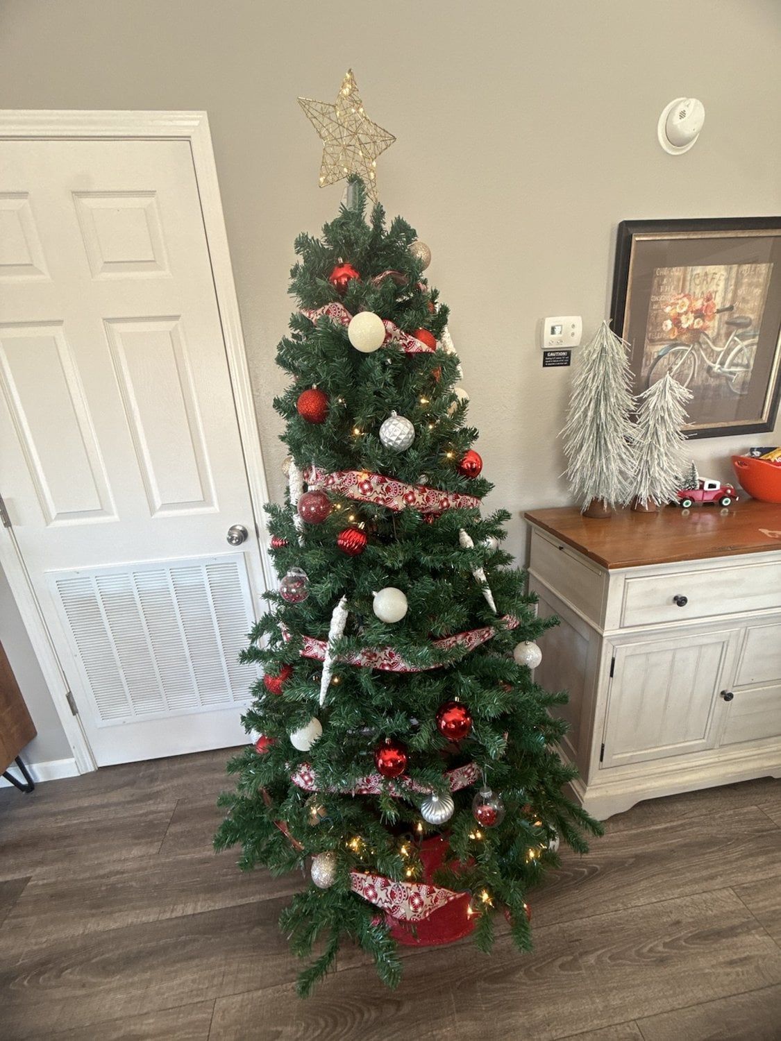 A Christmas tree with ornaments and lights, a white door, and a small decorative tree on a dresser.