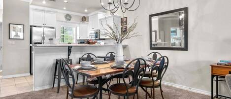 Comedor con mesa de madera, seis sillas, lámpara de araña y cocina al fondo.