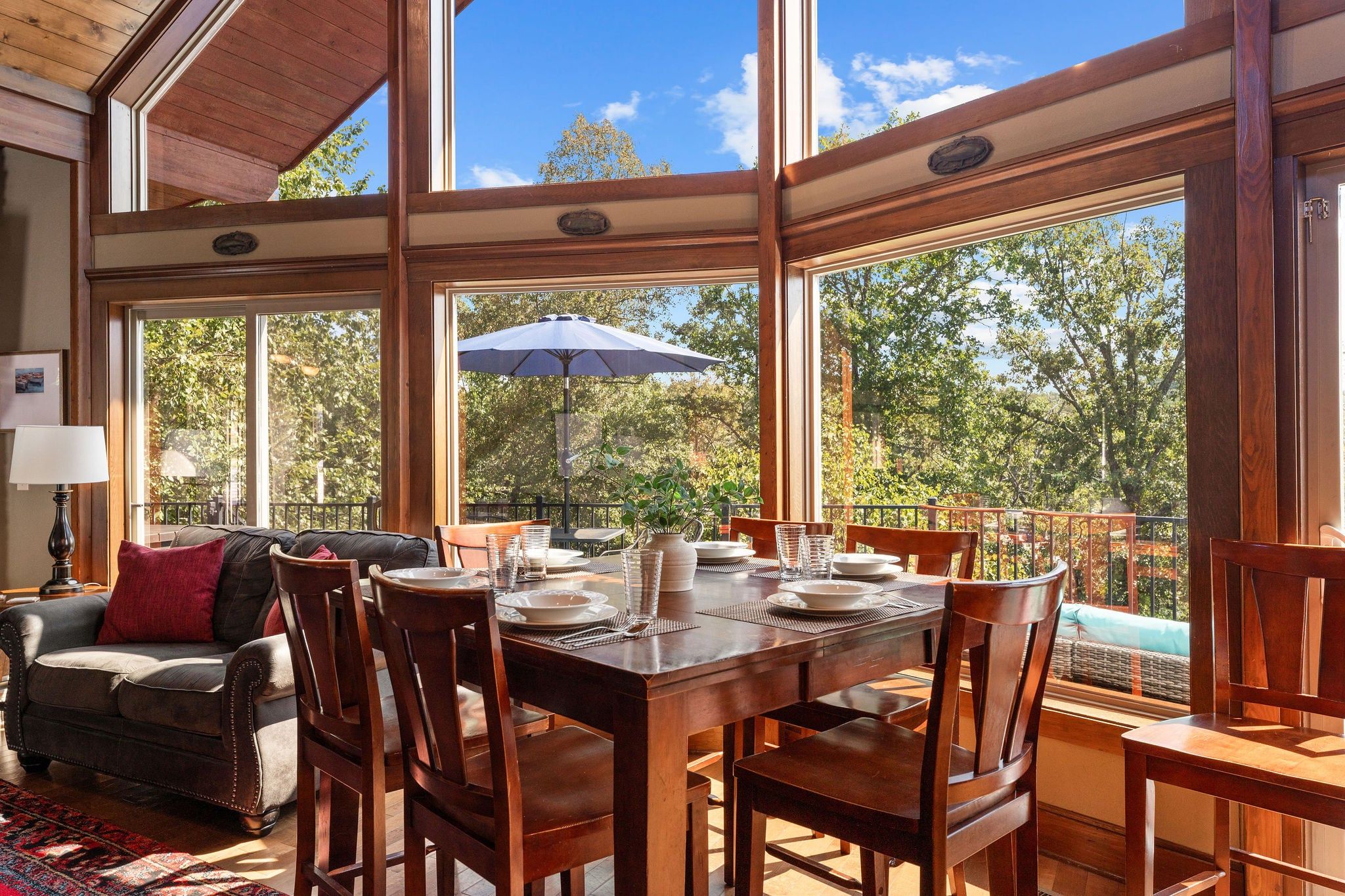A dining area with a wooden table set for four, surrounded by chairs, and large windows offering a view of trees and sky.