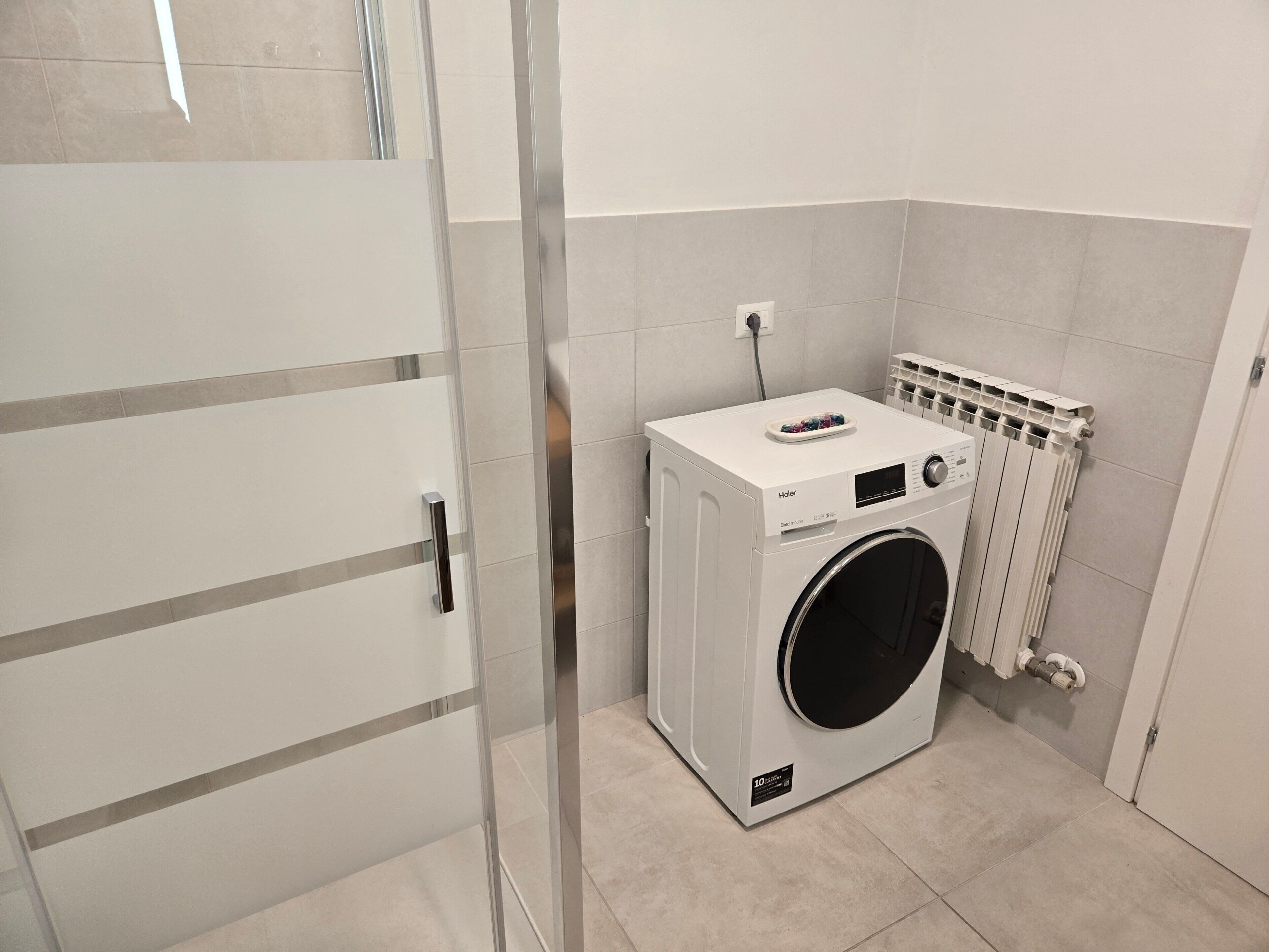 A compact laundry room with a front-loading washing machine, a wall-mounted radiator, and a door with a handle.