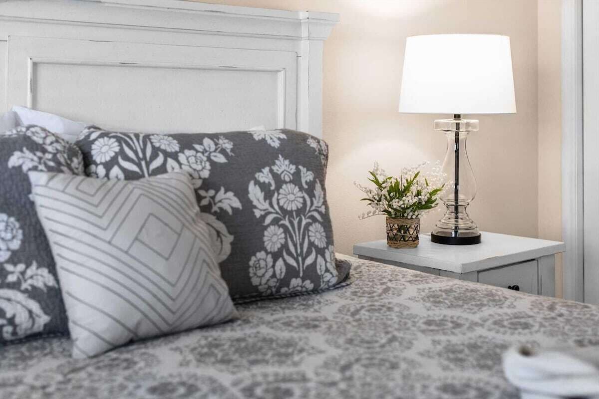 A neatly made bed with patterned pillows, a bedside table with a lamp and a small plant, and a white headboard.