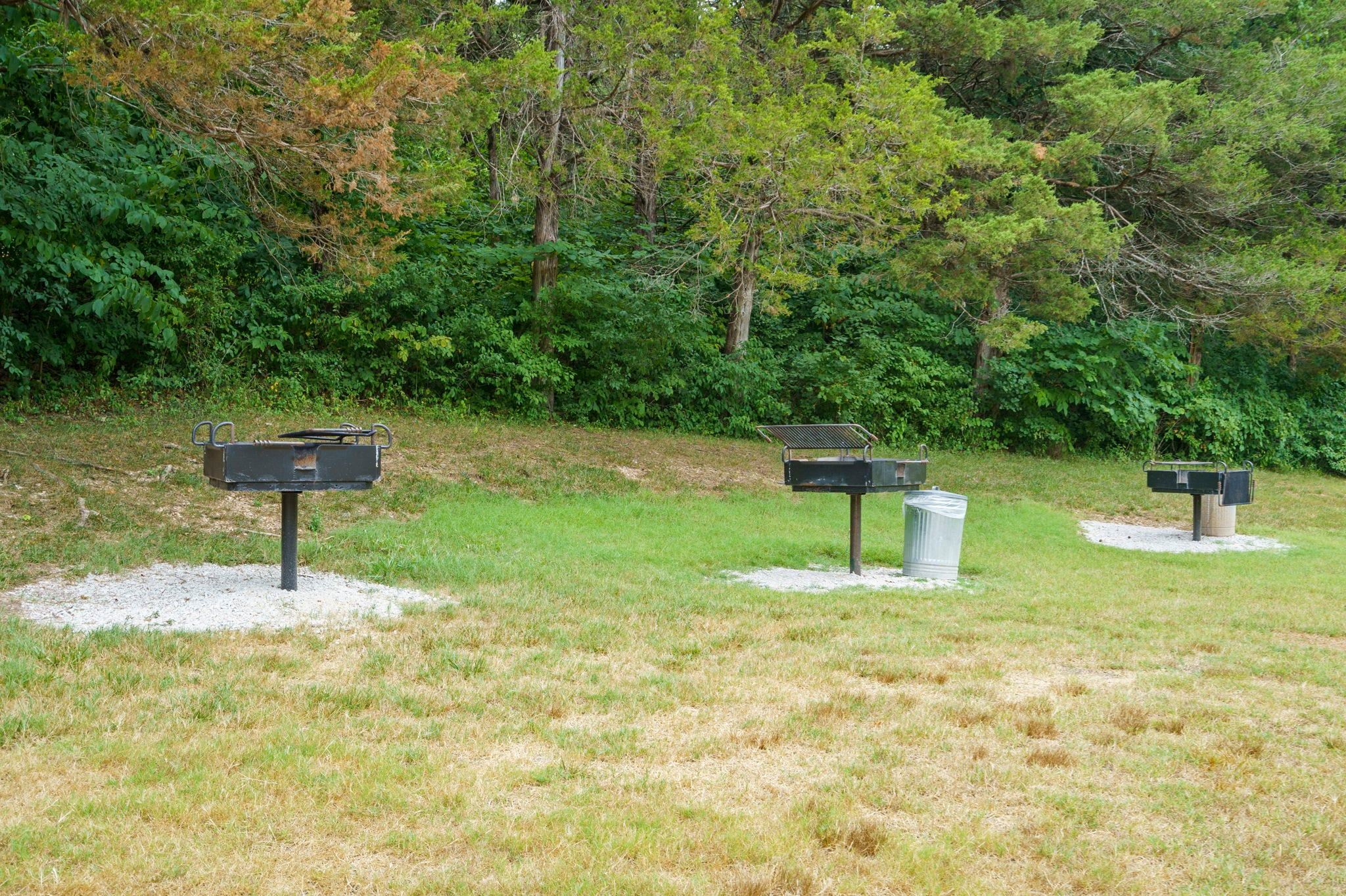 Three outdoor barbecue grills with metal grates, set on gravel bases, surrounded by grass and trees.