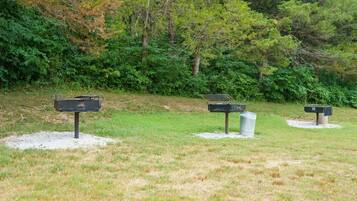 Three outdoor barbecue grills with metal grates, set on gravel bases, surrounded by grass and trees.