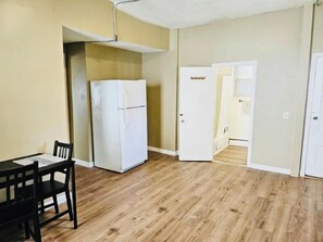 Quarto com piso de madeira, geladeira branca, mesa de jantar com cadeiras e porta que dá para outro cômodo.