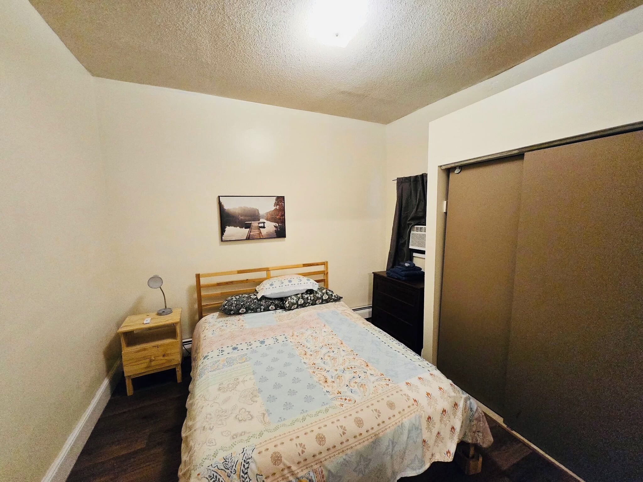 A bedroom with a bed, a nightstand, a desk, a picture on the wall, and a wardrobe.