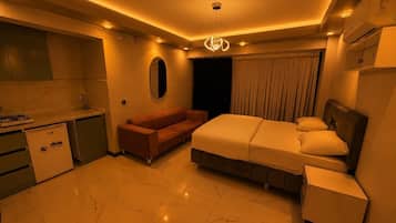 A modern hotel room with a kitchen area, a sofa, a bed with a headboard, and a small bedside table.
