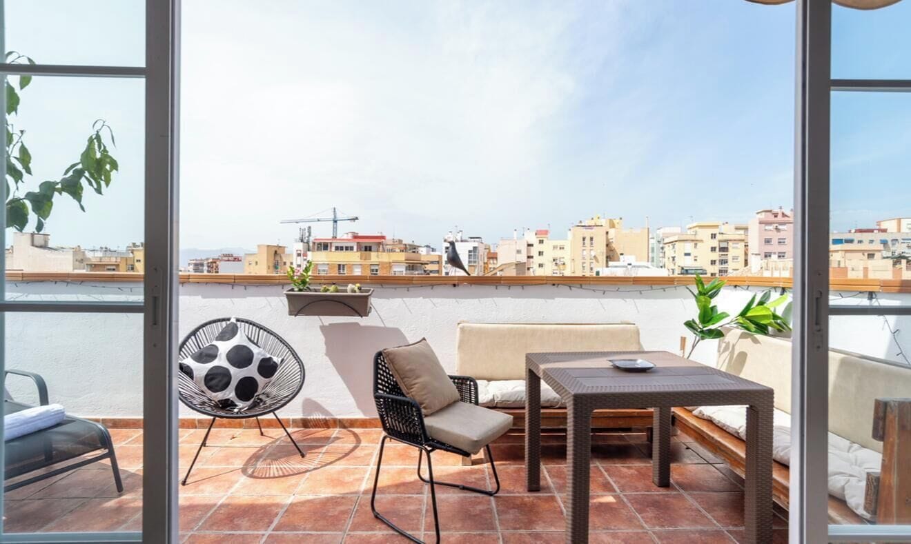 A balcony with a sofa, a chair, a table, and a view of the cityscape.