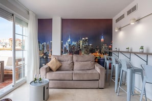 A modern living room with a cityscape mural, a sofa, a small round table with a bottle, and a bar area with stools. - New GR3 Penthouse Apartment in Downtown Málaga with a Cozy Terrace (Málaga)