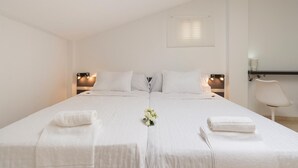 Uma cama de casal com lençóis brancos, duas mesas de cabeceira com luminárias, uma cadeira e uma pequena mesa com abajur.