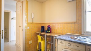 Uma cozinha compacta com máquina de lavar, uma cadeira amarela e uma pequena mesa com cafeteira.