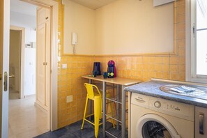 A compact kitchen with a washing machine, a yellow chair, and a small table with a coffee maker. - New GR3 Penthouse Apartment in Downtown Málaga with a Cozy Terrace (Málaga)
