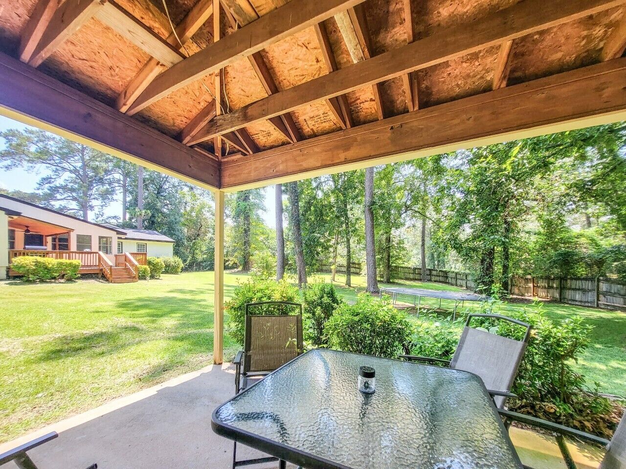 A covered patio with a table and chairs, surrounded by a grassy yard and trees.