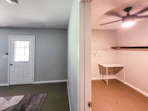 Quarto com porta, ventilador de teto e uma mesa branca encostada na parede.