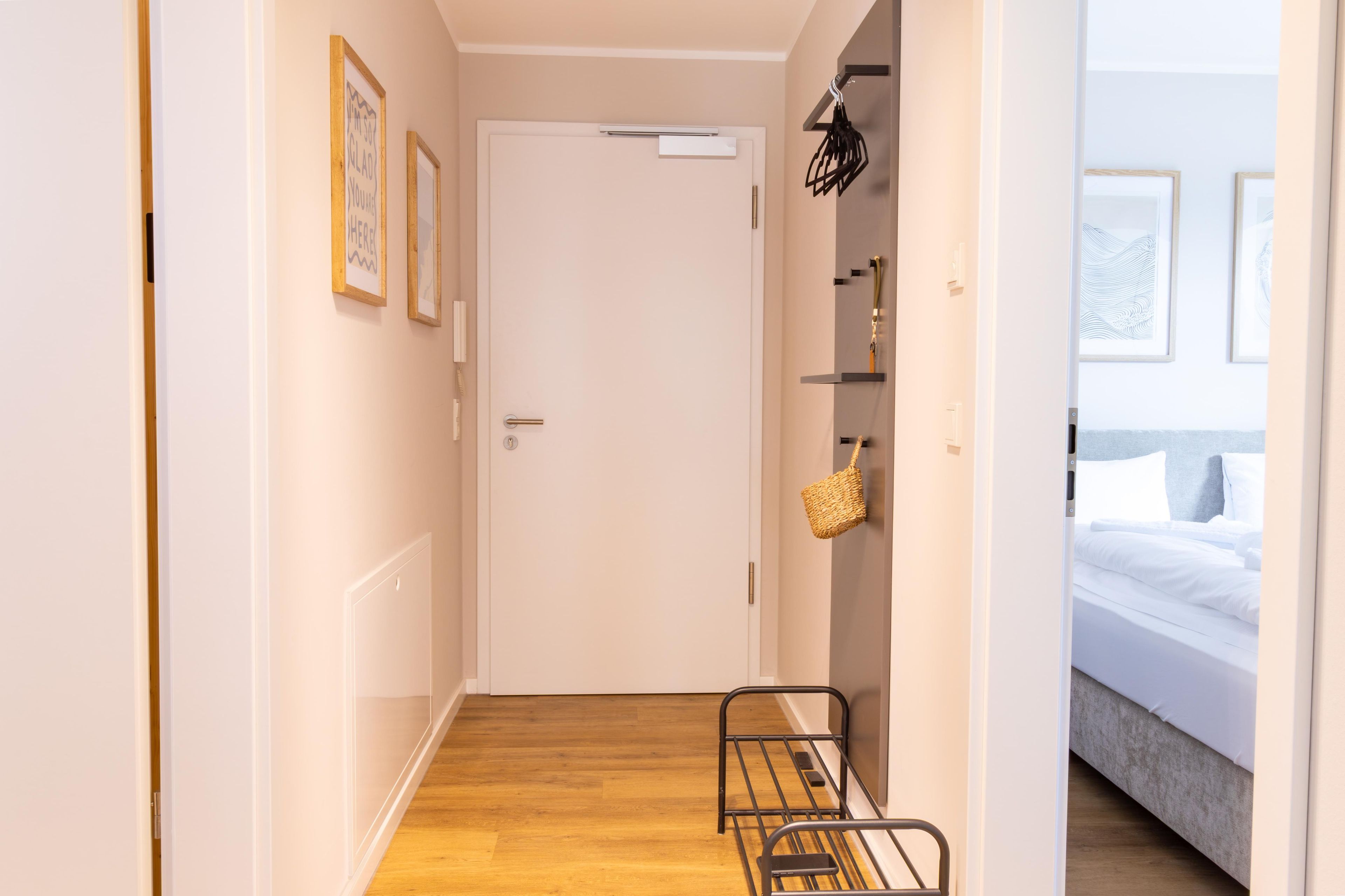 Un pasillo con piso de madera, una puerta blanca y una cama con ropa blanca en la habitación contigua.