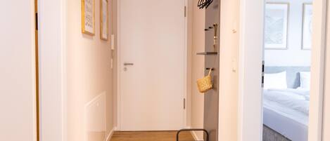 Un pasillo con piso de madera, una puerta blanca y una cama con ropa blanca en la habitación contigua.
