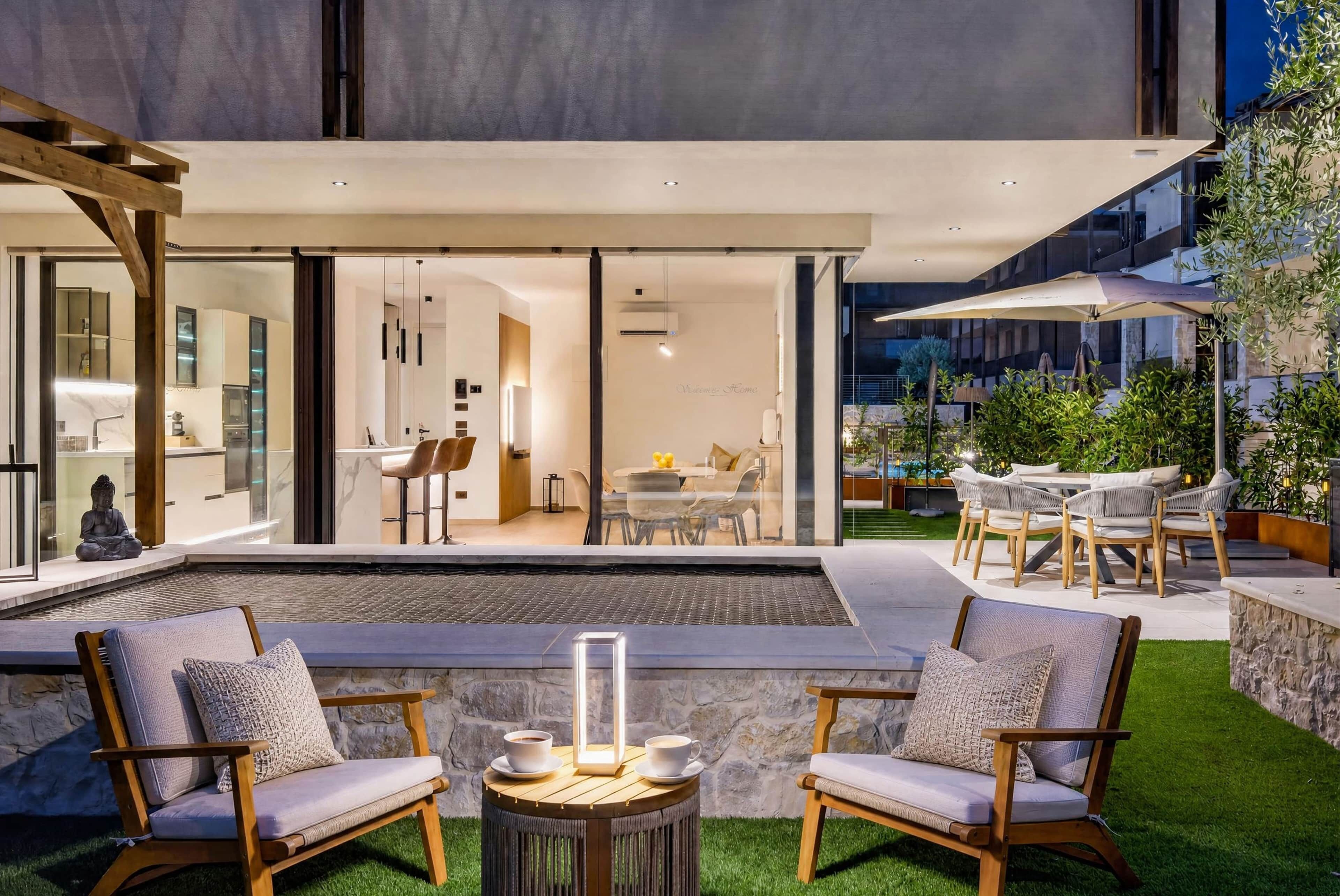 A modern outdoor seating area with two chairs and a table, a small pool, and a view of the interior of a contemporary house.
