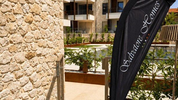 A stone wall, a flag with 'Salvatore Homes' text, and a building with balconies in the background. - NEW: Casa Serenia - Residence Corte Carden 2 (Bardolino)