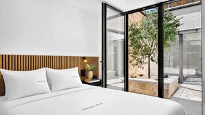 A modern bedroom with a large bed, a wooden headboard, a bedside table with a lamp, and a view of a garden through glass doors. - NEW: Casa Serenia - Residence Corte Carden 2 (Bardolino)