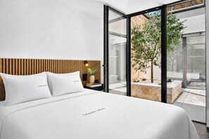 A modern bedroom with a large bed, a wooden headboard, a bedside table with a lamp, and a view of a garden through glass doors. - NEW: Casa Serenia - Residence Corte Carden 2 (Bardolino)